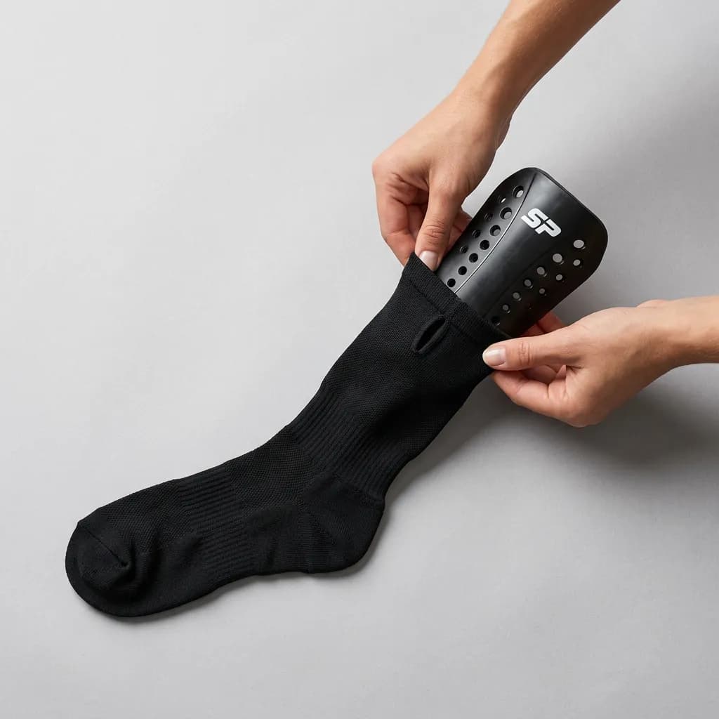 Händer som för in ett svart SP-benskydd i fickan på en svart SOCKPOCKET-strumpa